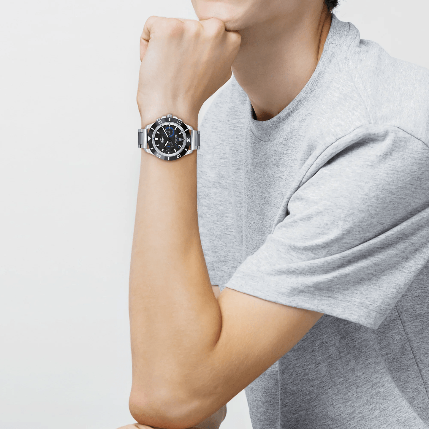 A person in a gray t-shirt, resting their chin on their hand, is wearing a Lacoste Toronga Dual Time Black Dial Silver Bracelet Watch featuring quartz movement and a sleek black face.