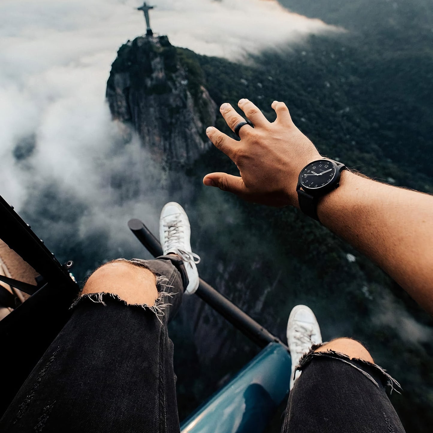 Wearing ripped jeans and an MVMT Field Gunmetal Adventure watch, a person reaches from a helicopter near Christ the Redeemer.