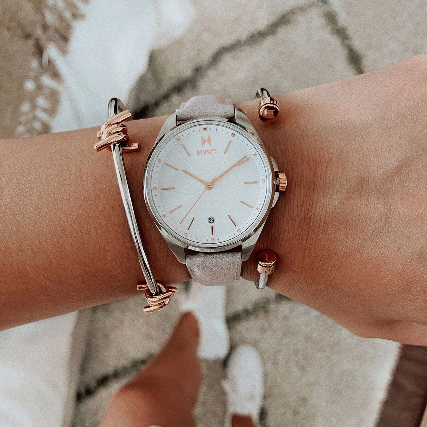 Wrist with a MVMT Ladies Coronada Malibu Date watch, stylish bracelet, and casual beige shoes on a textured background.
