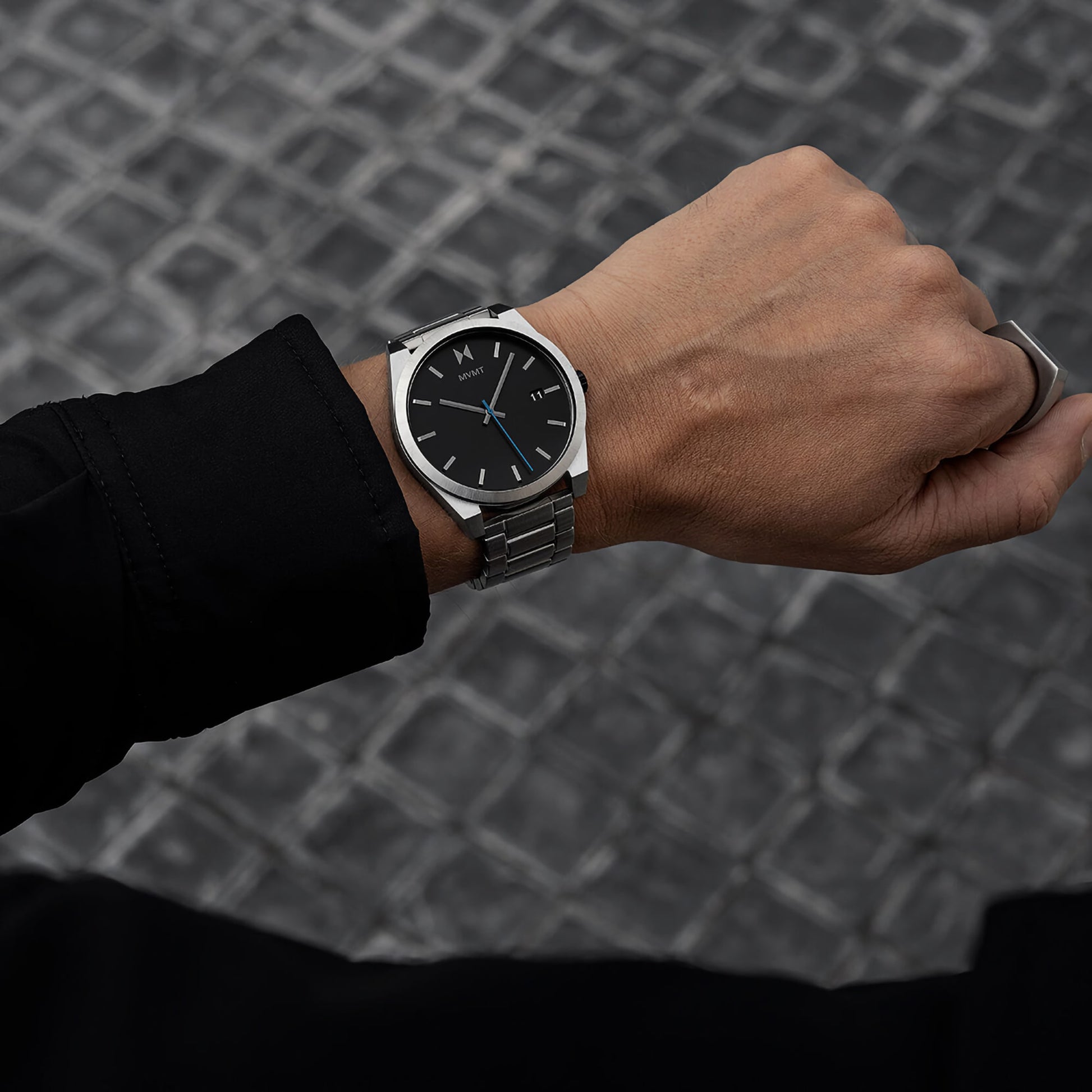 Close-up of the MVMT Element Ion Silver men's watch with a black face and silver band against a cobblestone backdrop.