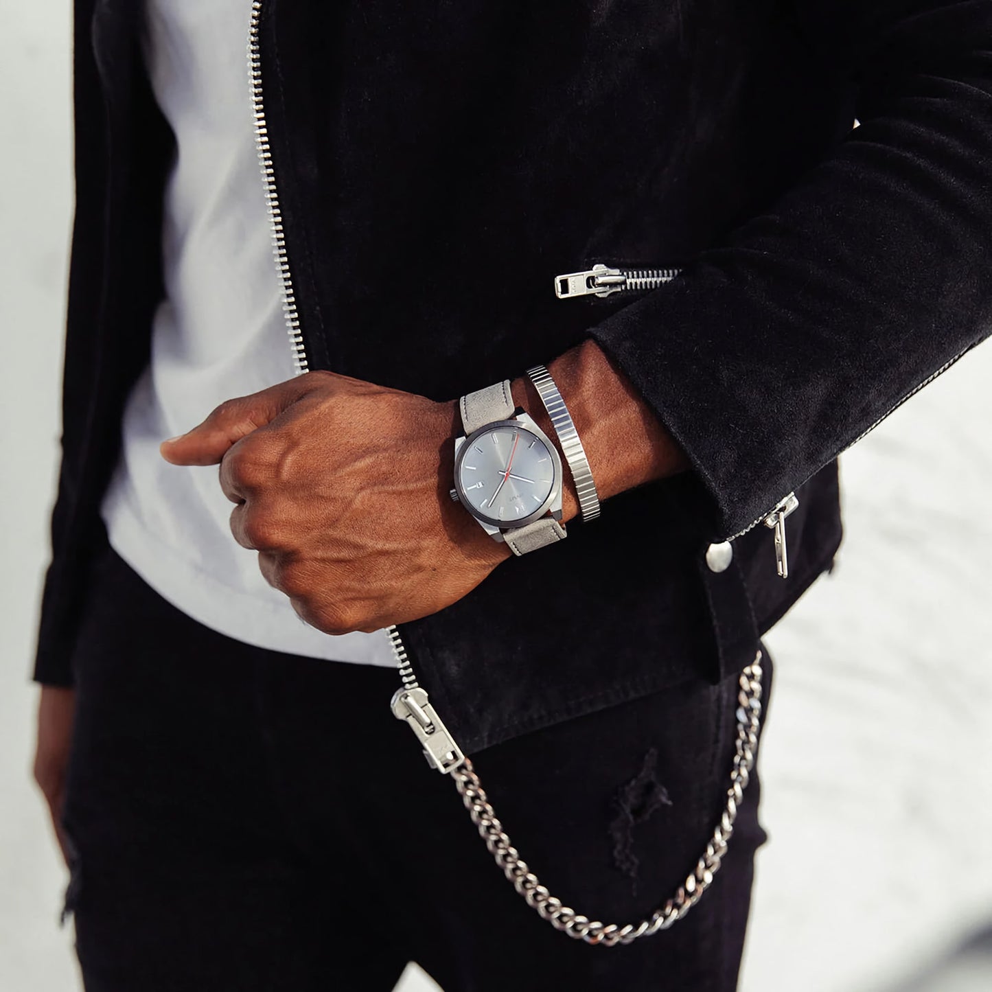 A person in a black jacket and white shirt wearing an MVMT Element Radium Grey watch and bracelet, with a chain hanging from jeans.