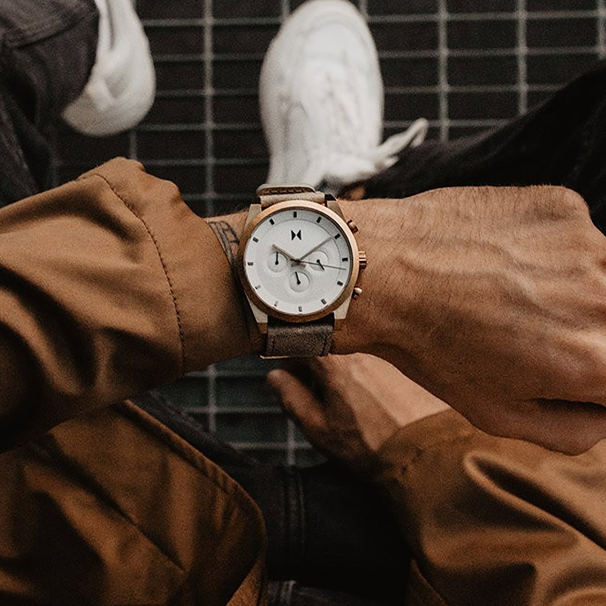 Close-up of a person wearing the MVMT Element Chrono Bronze Ore watch, dressed in a brown jacket and white sneakers.