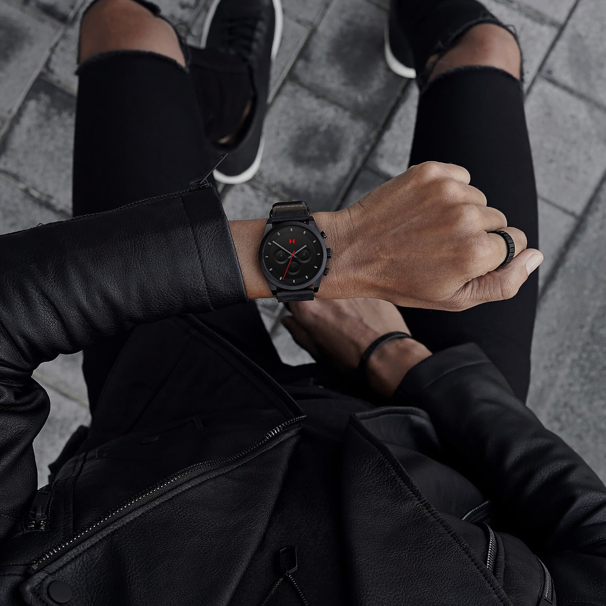 Person in a black leather jacket and pants flaunting an MVMT Element Chrono Ember Black watch while seated on a pavement.