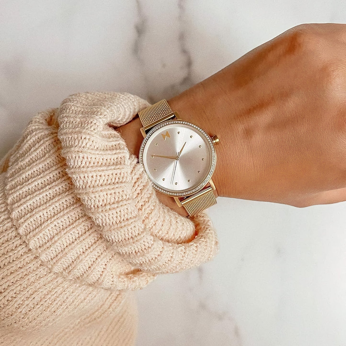 Wrist with an MVMT Dot Crystals Ladies watch and a beige sweater sleeve on a marble background.