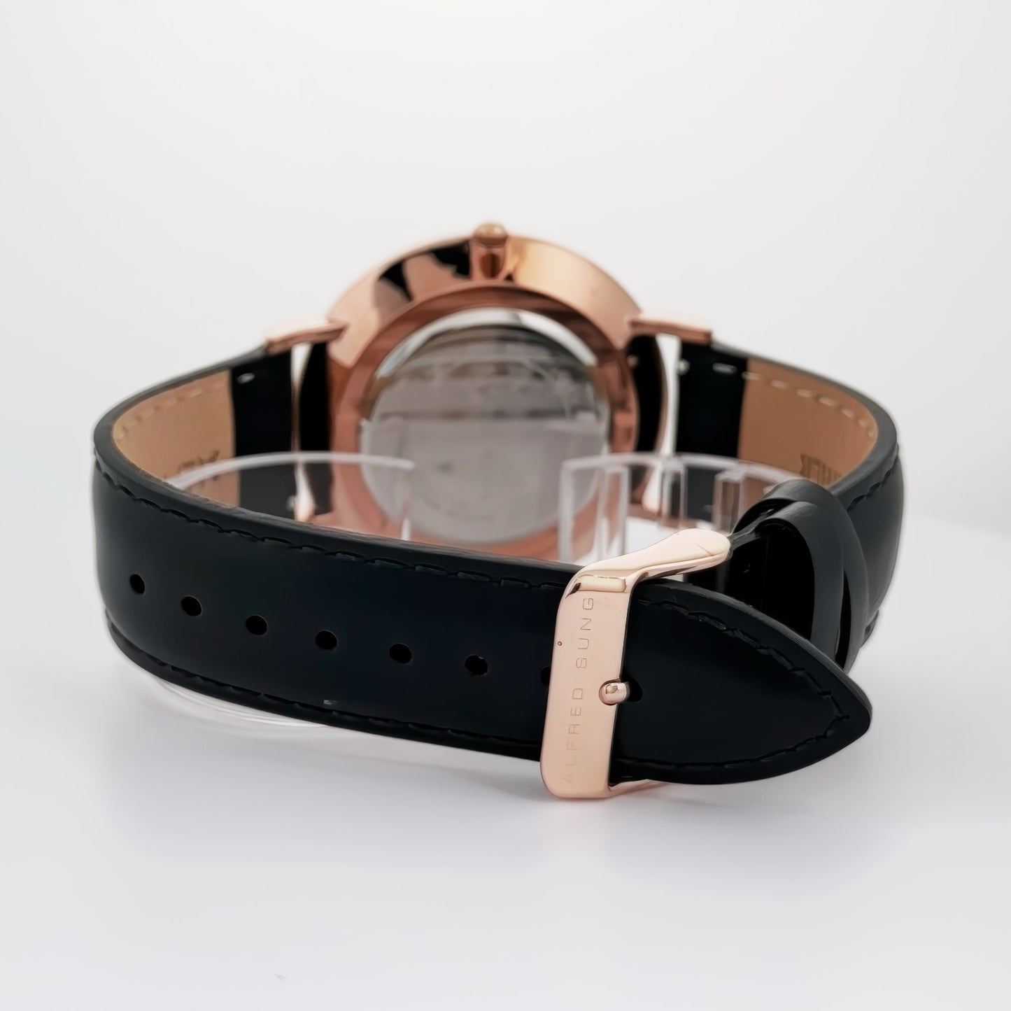 Back view of an Alfred Sung Metropolitan Date Black Dial watch with black leather strap and rose gold buckle on white surface.