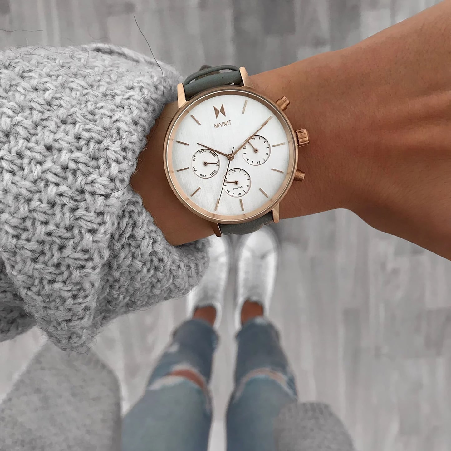 Person wearing an MVMT Nova Dorado watch, dressed in a gray sweater and ripped jeans.