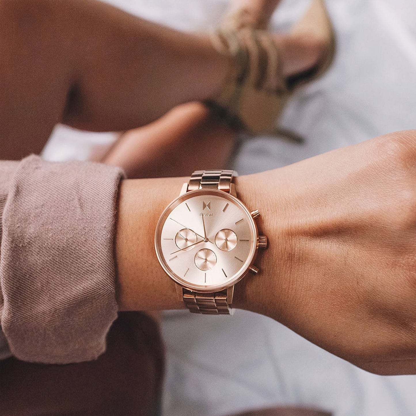 Wrist with MVMT Nova Leo Rose Ladies watch, Japanese quartz, in focus; blurred legs in background.