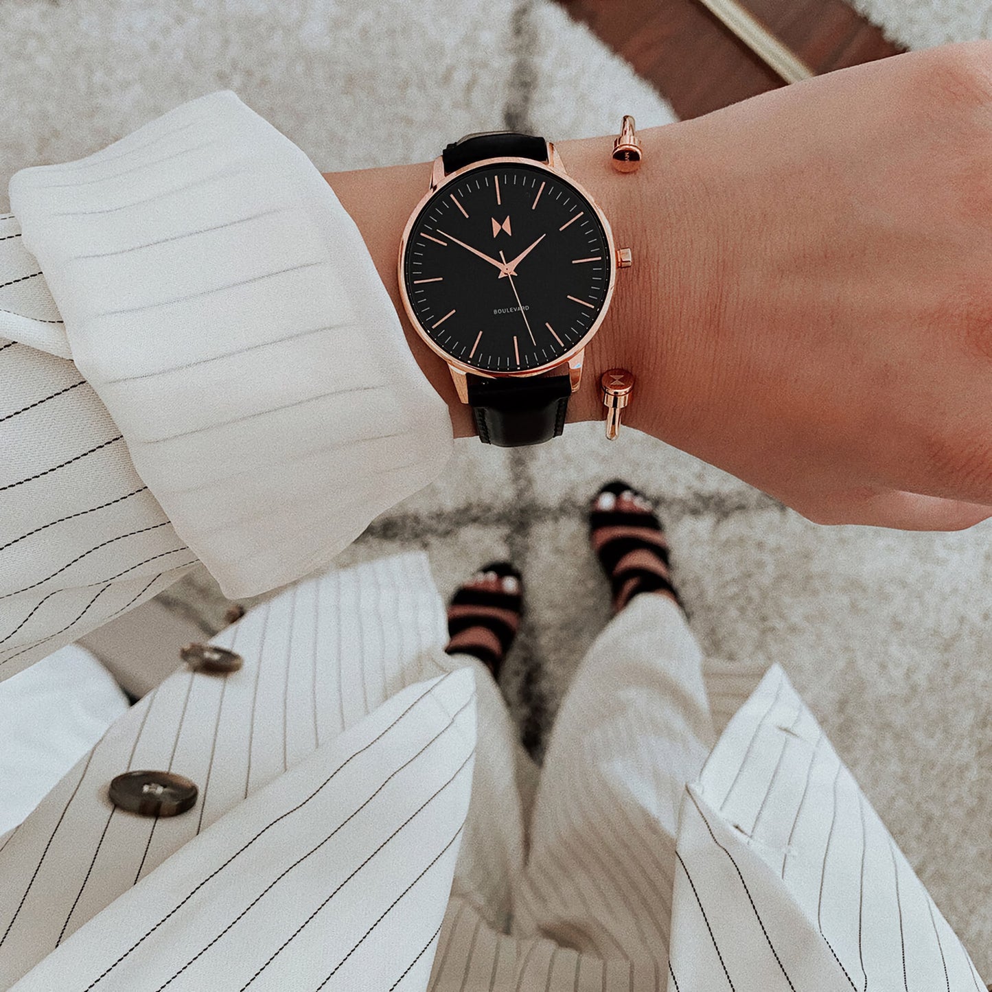 Close-up of a wrist accentuated by an MVMT Boulevard Santa Monica Black watch paired with a gold bracelet and pinstriped outfit.
