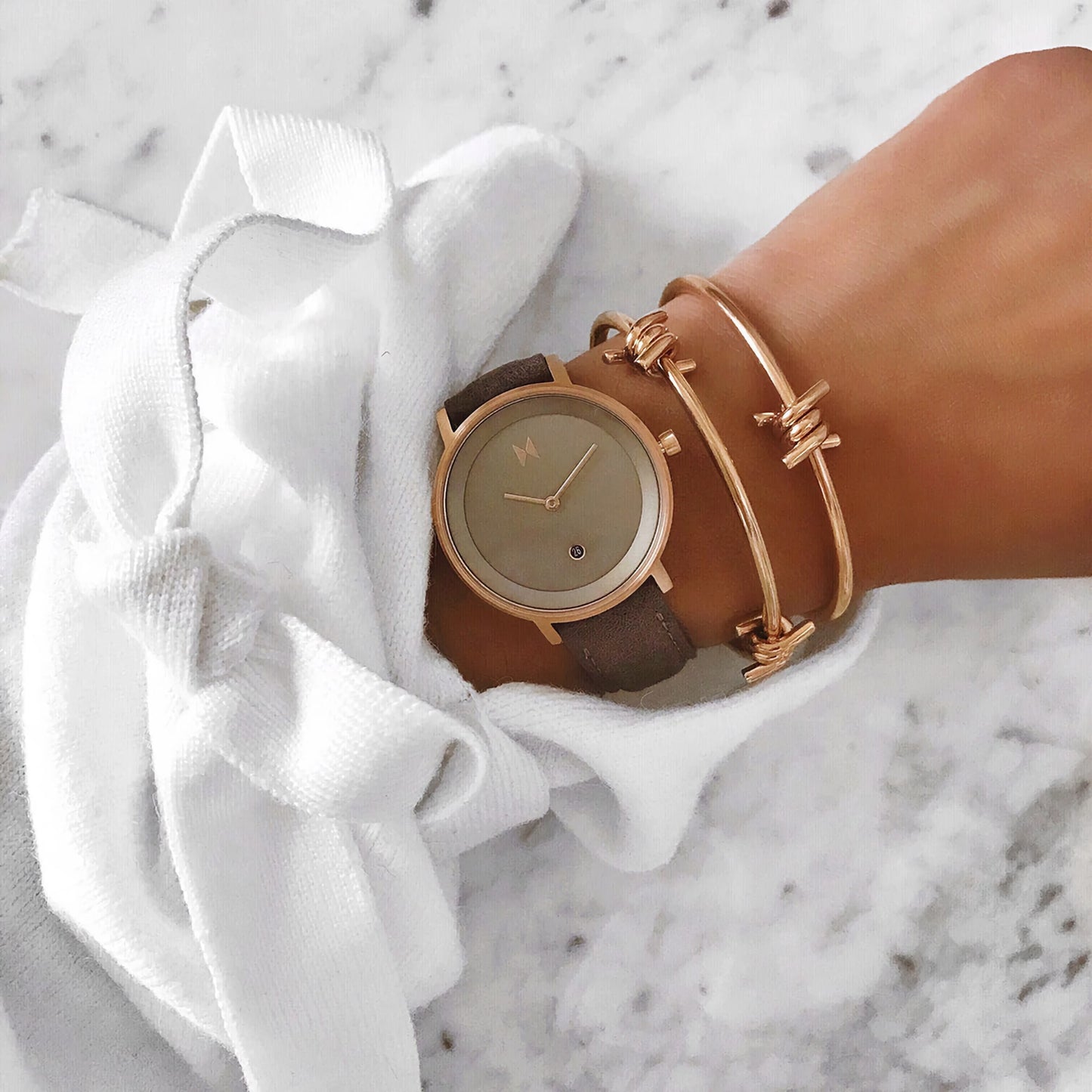 An MVMT Signature II Ashen Taupe watch with bracelets on a wrist, resting on a white cloth over a marble surface.