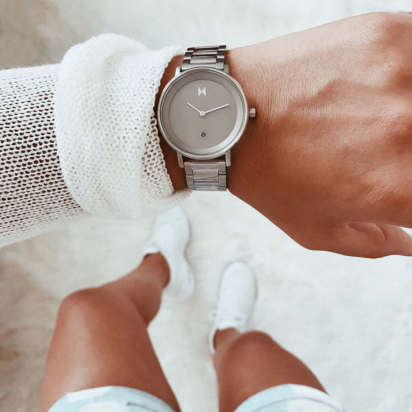 A person in a white sweater and shorts wears an MVMT Signature II Cloud Silver Ladies watch on a light carpet with white sneakers.