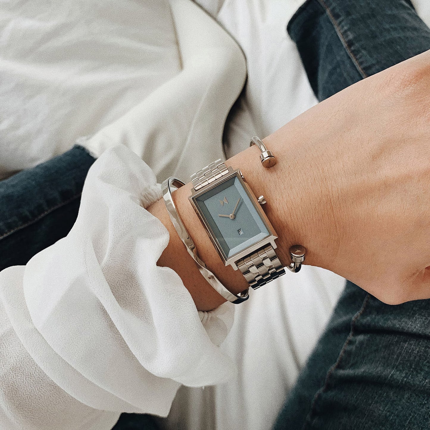 A person wearing an MVMT Signature Square Skylar Blue watch and bracelets, in a white blouse and jeans.