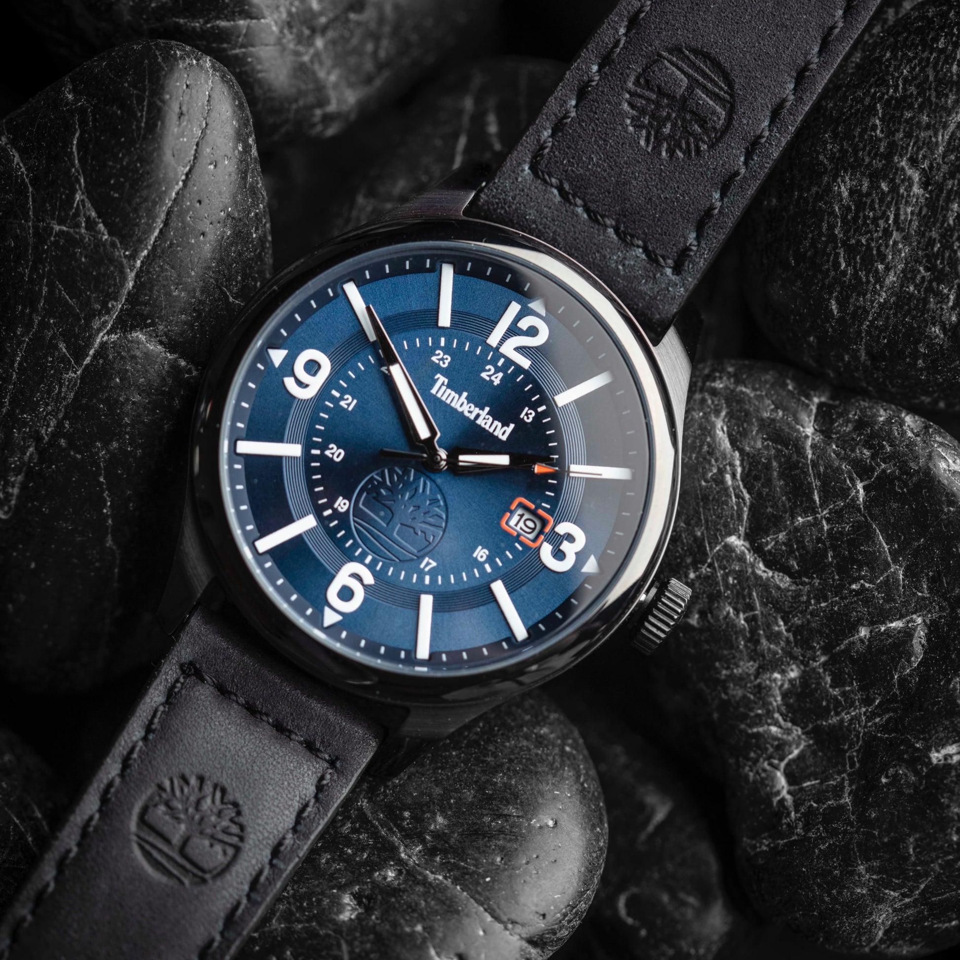 A Timberland Blake Blue Date watch with a black leather strap rests elegantly on black stones.