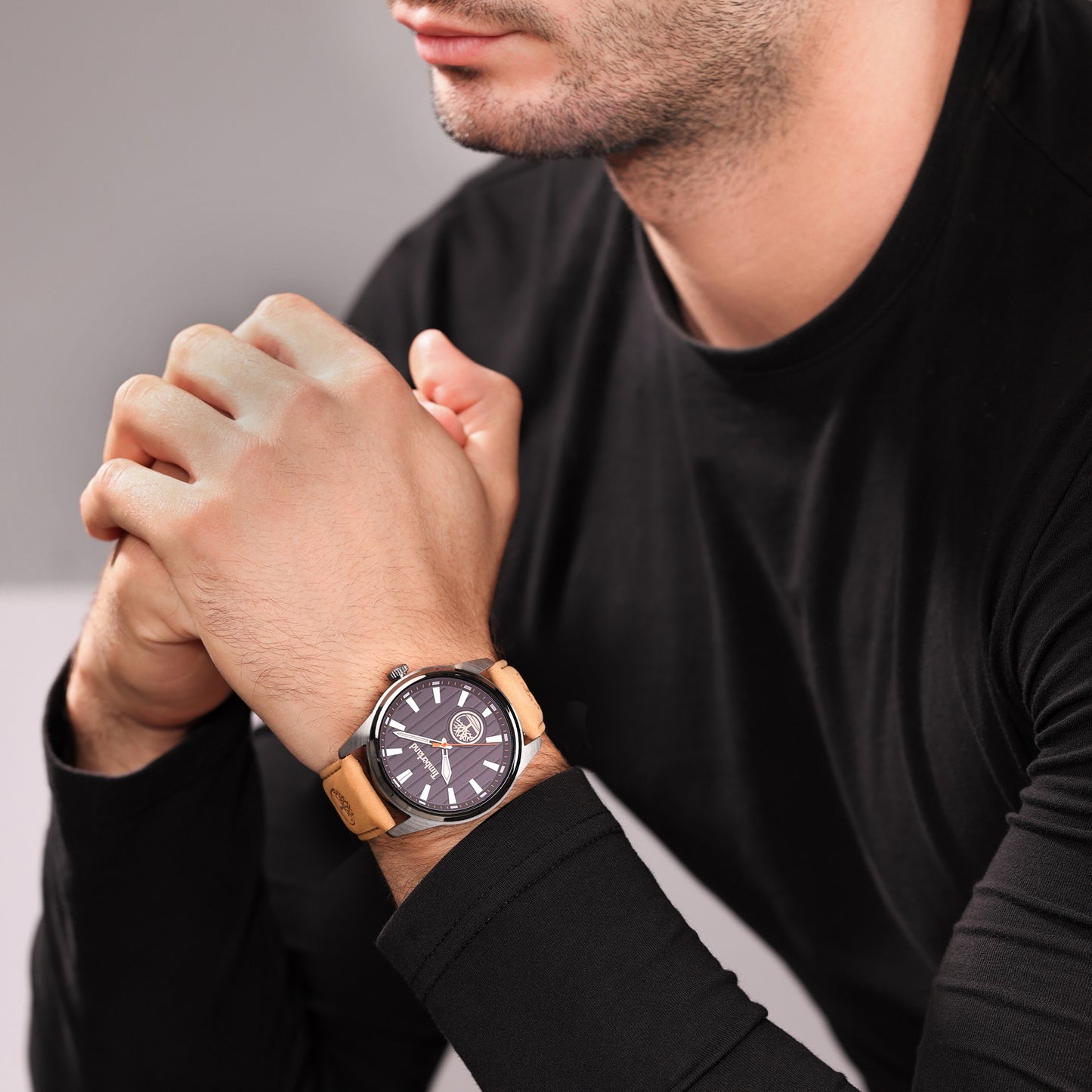 Man in black shirt wearing Timberland Northbridge watch with a brown leather strap, hands clasped together.