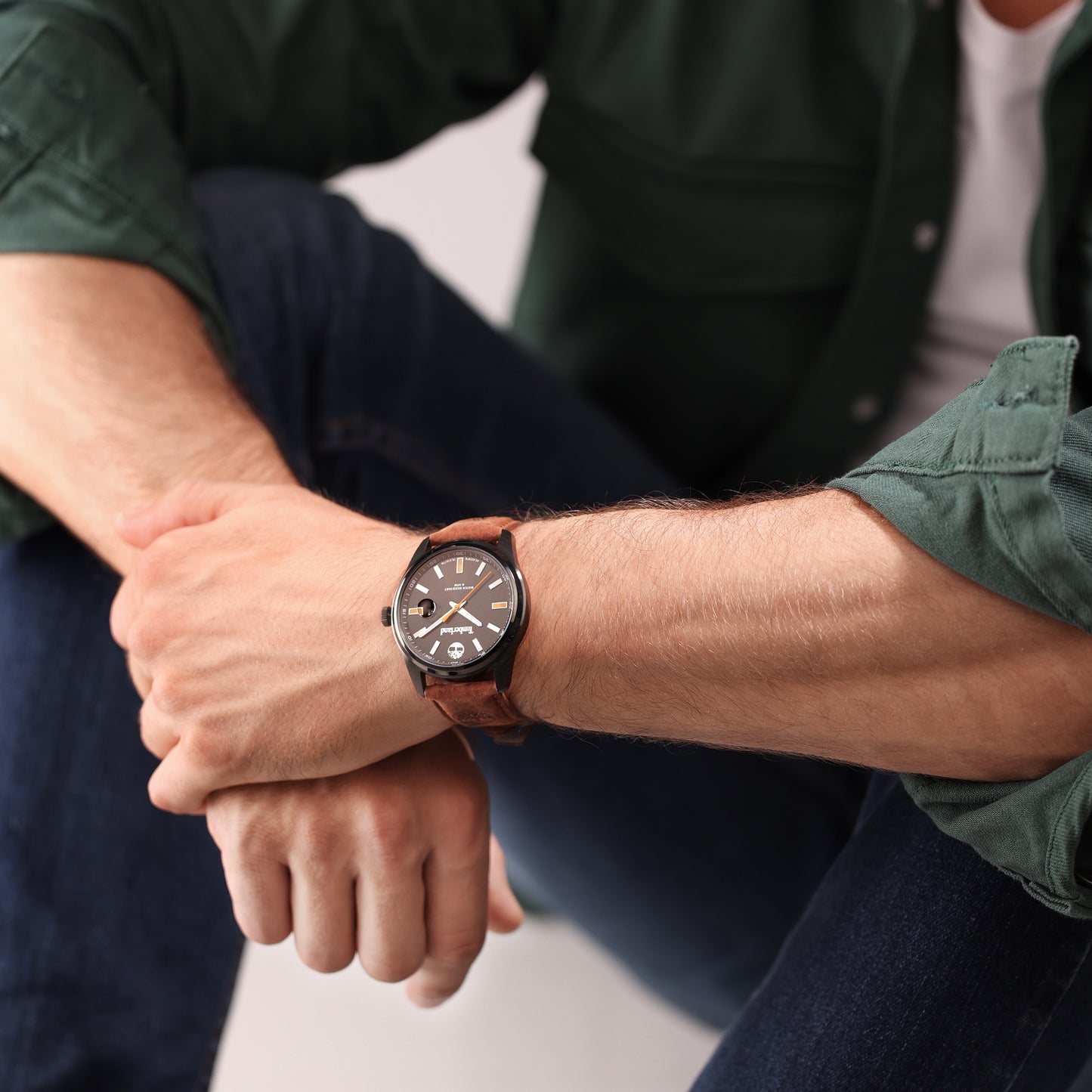 Person in green shirt and jeans wearing a Timberland Orford watch, sitting with elbows on knees.