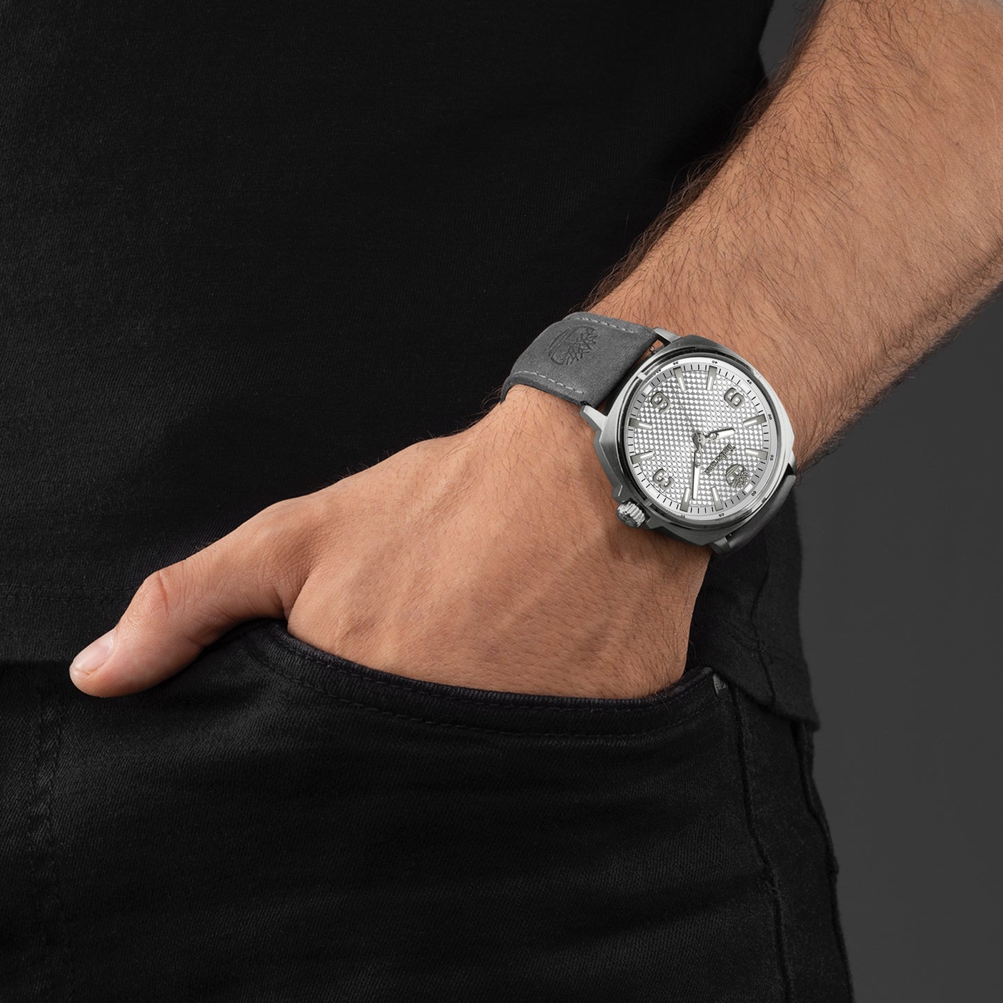 Person wearing black shirt and pants, Timberland Bailard watch, hand in pocket against a gray background.