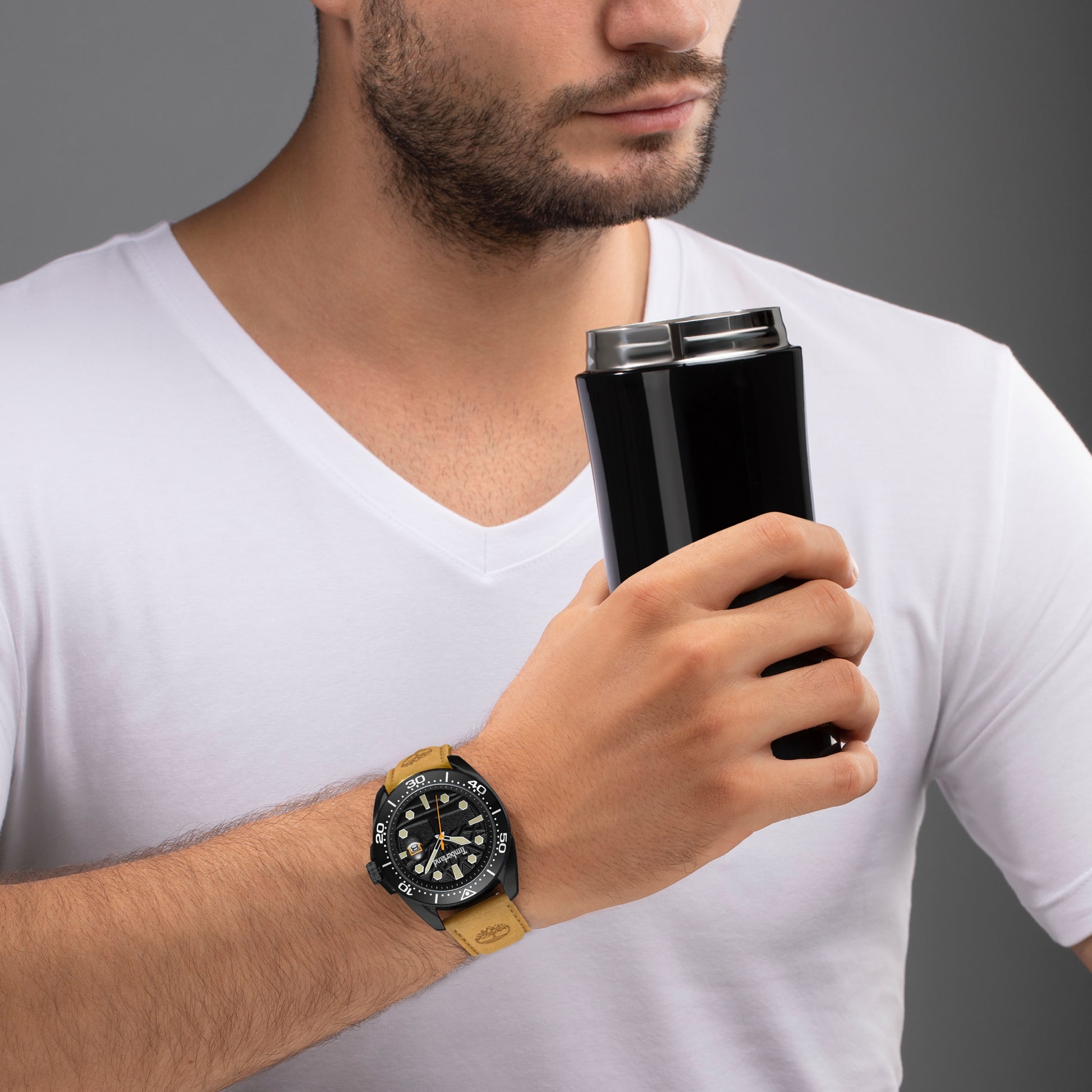 Man in a white shirt holding a black travel mug, wearing a Timberland Carrigan watch, gray background.