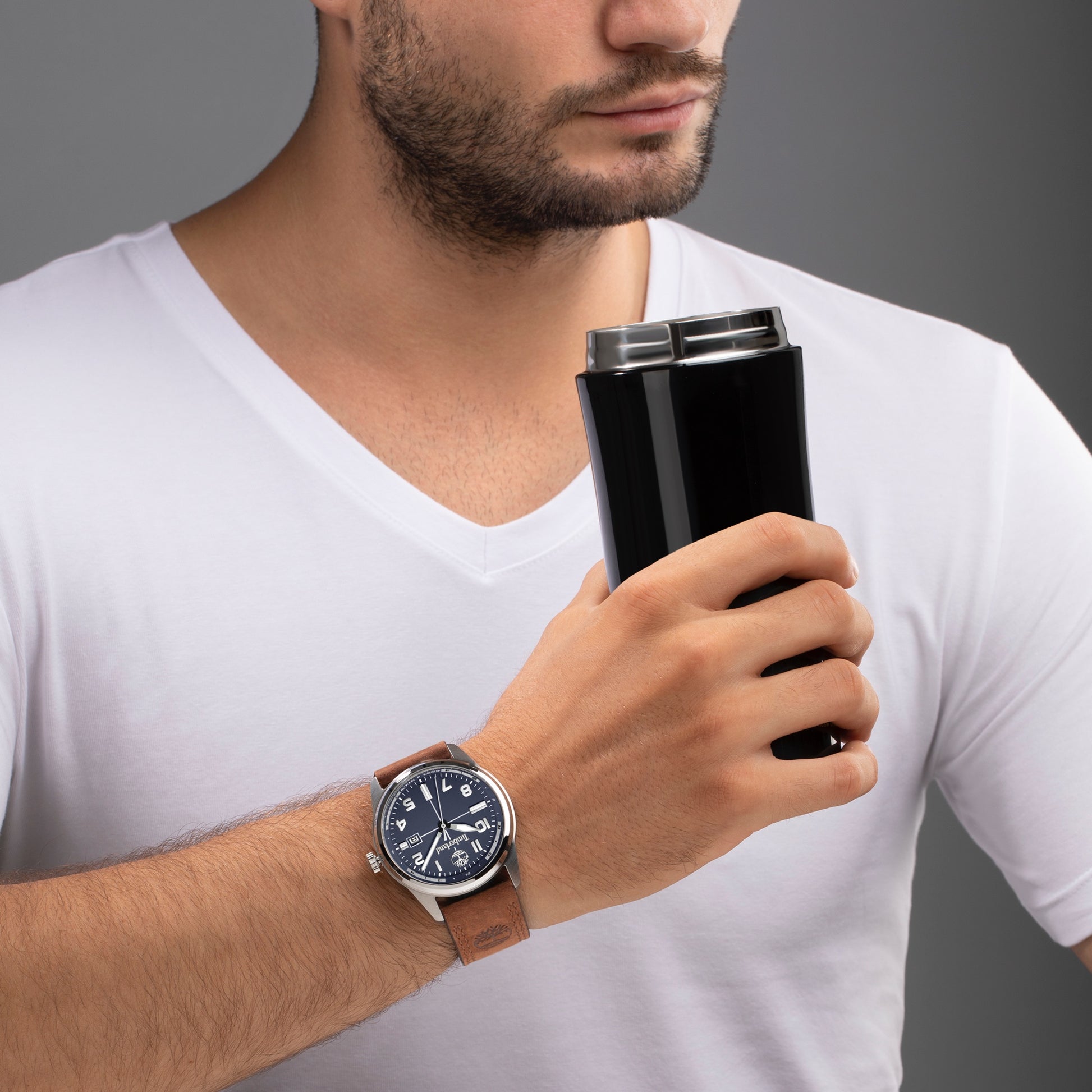 Man in a white shirt holding a black travel mug, wearing a navy Timberland Sherbrook Men's Watch with a brown leather strap.
