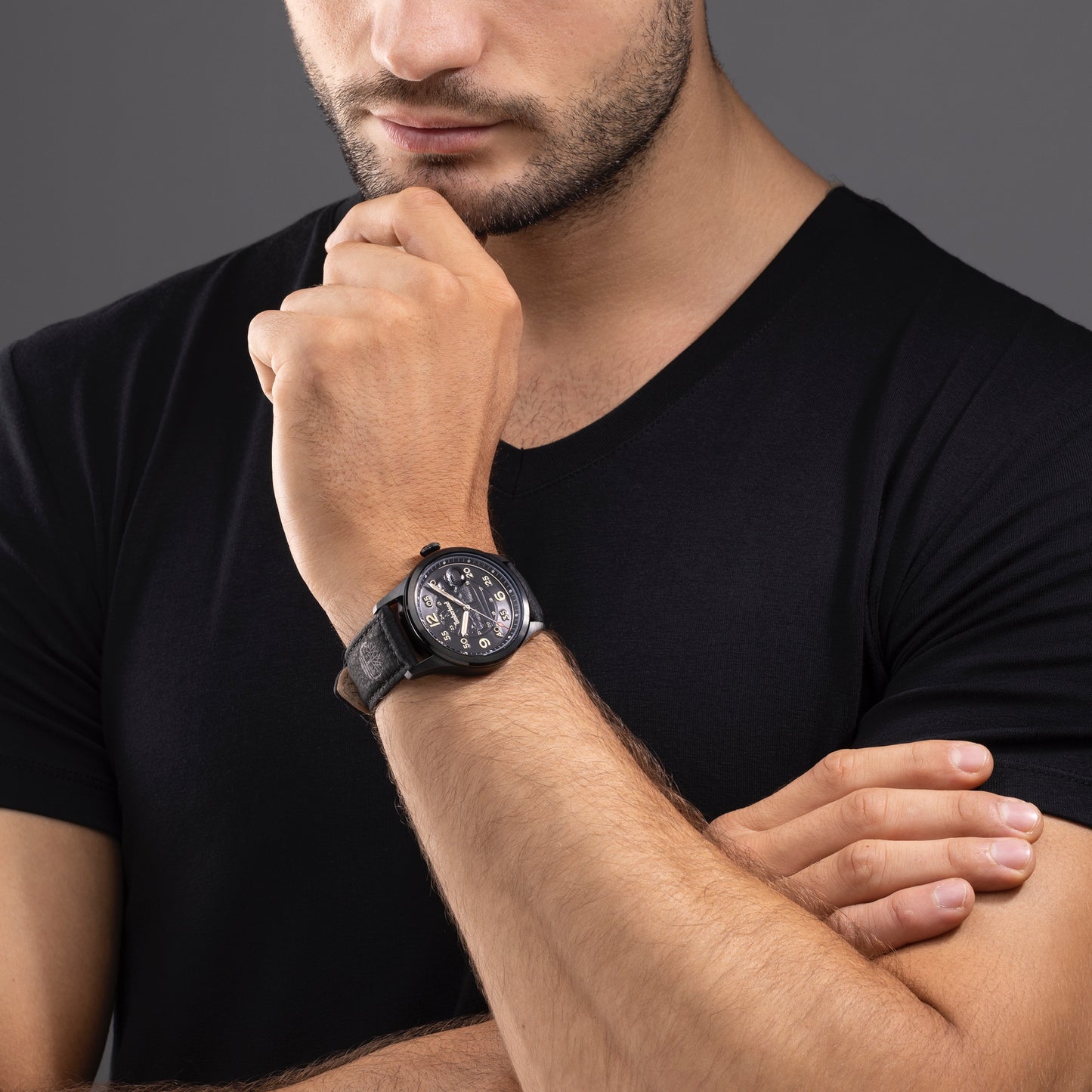 Man in black T-shirt wearing a Timberland Cornwall watch, holding his chin thoughtfully against a gray background.