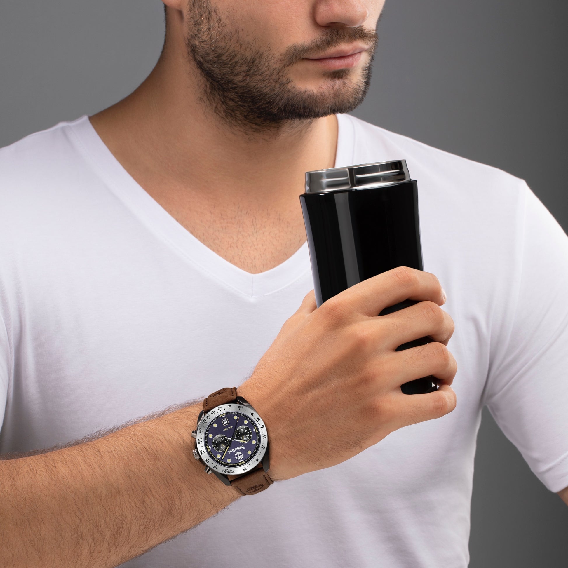 A man in a white shirt holds a black travel mug, wearing a Timberland Carrigan watch with quartz movement on his wrist.