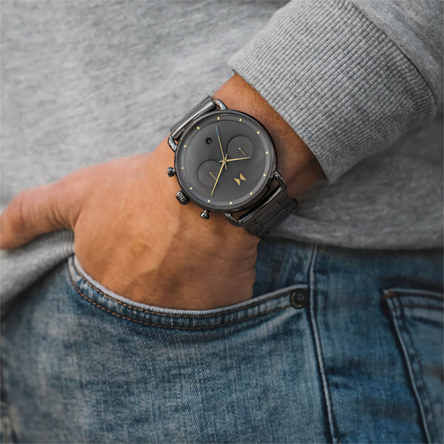 Close-up of a person wearing the MVMT Blacktop Honey Smoke Grey Dial watch, featuring a grey IP bracelet and stylish gold accents, with their hand in the pocket of blue jeans.