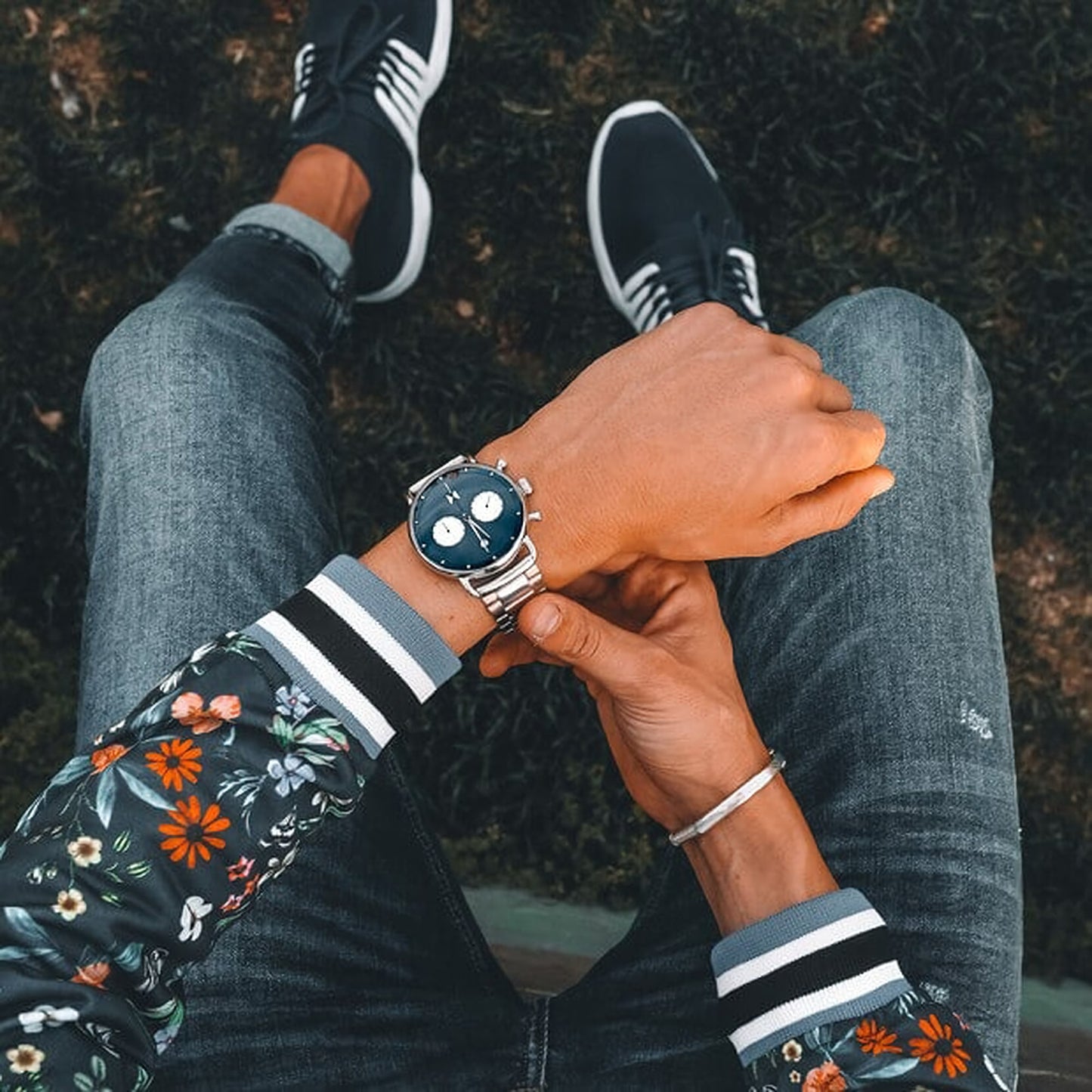 A person in a floral jacket and sneakers is checking the MVMT Blacktop Blue Dial Stainless Steel Bracelet Men's Watch equipped with a Miyota 6S21 movement while sitting and seen from above.